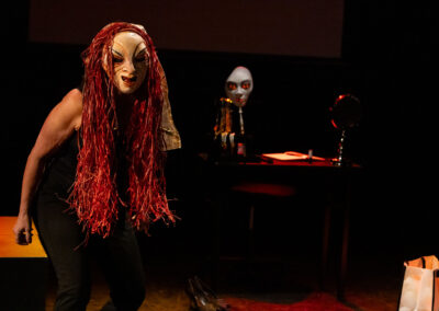 A performer in a theater wears a striking mask with long, red hair-like strands. They are in a dark set with a table in the background, holding another mask and some props. The scene conveys an intense and dramatic atmosphere.