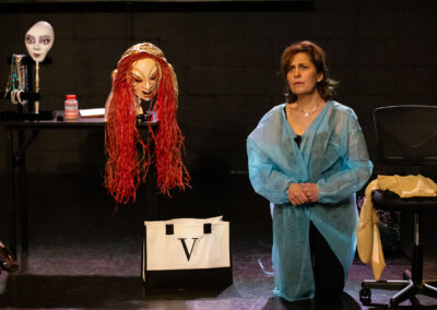 A woman wearing a blue protective gown kneels on the floor next to a white and black bag. Behind her is a table with two masks, one with red hair and the other white. The scene appears to be set in a dimly lit room with black walls and an office chair nearby.