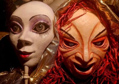 Two expressive masks side by side in close-up. The left mask is white with big, round, purple-shadowed eyes and red lips, draped in metallic fabric. The right mask has a reddish hue with exaggerated features and red, tangled hair-like adornment.