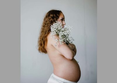 Photograph of profile of pregnant woman, with a half slip and a bare torso, holding white flowers in front of her.