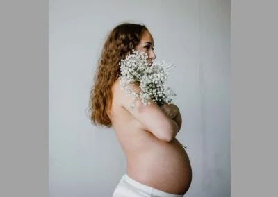 Photograph of profile of pregnant woman, with a half slip and a bare torso, holding white flowers in front of her.